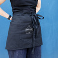 A black waist apron with a front pocket and a tied waist, held by a person against a blue background.
