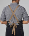 A denim bib apron with a cross-back design and a brown tie, worn by a person in a checked shirt.