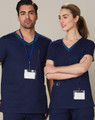 A navy scrub top with contrasting teal collar details, worn by a male and female model. Features pockets and identification badges.