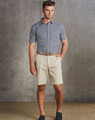 Men's chino shorts in beige, paired with a checked short-sleeve shirt and a brown belt. The model stands against a textured grey backdrop.