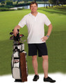 A man stands by a golf bag wearing navy chino shorts and a white polo shirt, with a lush green landscape in the background.