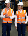 Two individuals wear bright orange Hi-Vis safety vests with reflective tapes and hard hats in a warehouse setting.