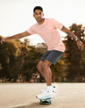 A man in a light pink premium cotton tee skates on a skateboard, wearing grey shorts and white sneakers.