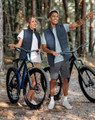 A pair of models wearing navy insulated puffer vests, standing beside bicycles in a forest setting.