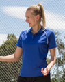 A woman in a blue polo shirt with white accents stands outdoors near a fence, smiling and gesturing.