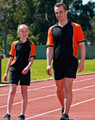 Men's TrueDry tri-colour short-sleeve pique polo in black and orange, paired with matching shorts, worn by two people on a track.