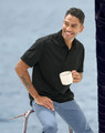 A man sits on a railing, wearing a black sustainable jacquard knit polo shirt and holding a white mug.