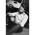 A man wearing a green button-up shirt sits on the ground, looking at his wrist, with a vintage car in the background.