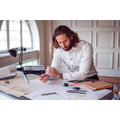 A man with long hair wearing a white button-up shirt, sitting at a desk with architectural plans and tools.