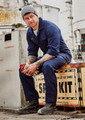 A man in a navy blue lightweight cotton drill overall, seated on a box outside, wearing a beanie and work boots.