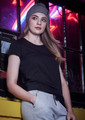 A woman in a black t-shirt and grey pants leans against a window, wearing a grey beanie. The background features neon lights.