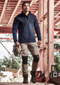 A man wearing a navy waterproof jacket stands on a construction site, holding a saw. He pairs it with beige pants and boots.