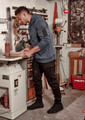 A man wearing a navy blue hoodie and black stretch jeans stands in a workshop, handling a piece of wood.
