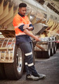 A man in a high-visibility orange and navy work shirt and dark stretch jeans leans against a truck.