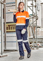 A woman in an orange and navy work shirt and matching pants stands on a construction site, holding wooden planks.