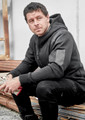 A black unisex zip front hoodie with multi-pockets, featuring a logo, worn by a seated man.