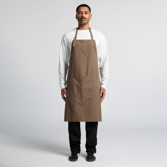 A man stands wearing a brown canvas apron over a white shirt, featuring a logo and front pockets.