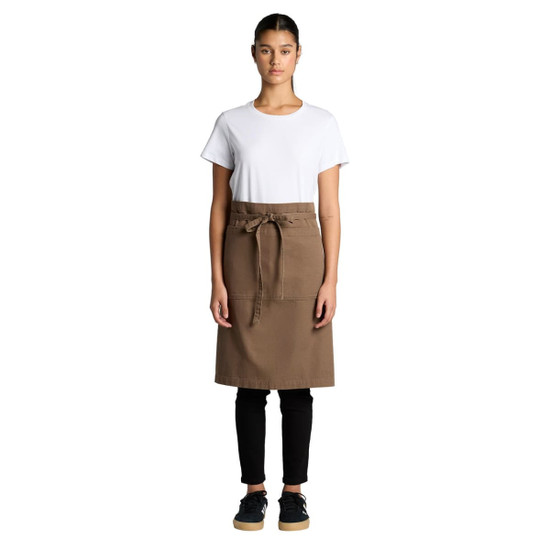 A woman wearing a brown canvas half apron over a white t-shirt and black pants, with a branding logo present.