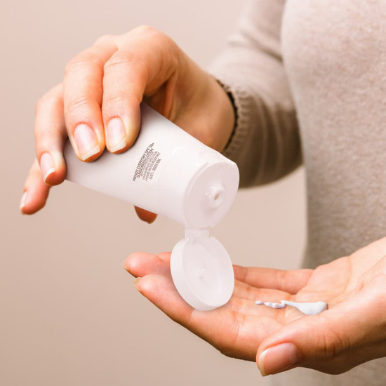 A white sunscreen tube being squeezed, dispensing a small amount of white cream onto a hand. The tube features a logo.