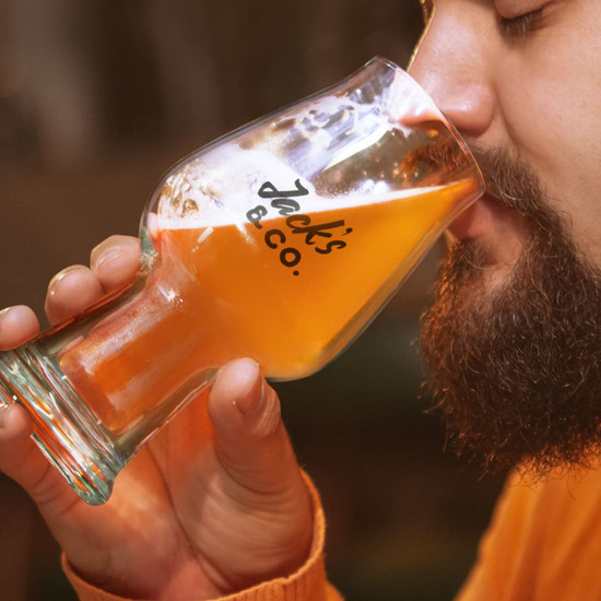 A beer glass with a logo, featuring a curved design, holds a golden beverage near a person's face.