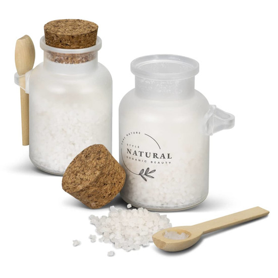 A glass jar containing white bath salts with a cork lid and a wooden spoon nearby. The jar has a simple branding design.