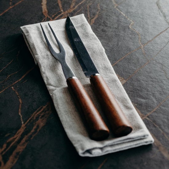 A cheese carving set featuring a knife and fork with wooden handles, positioned on a linen cloth.
