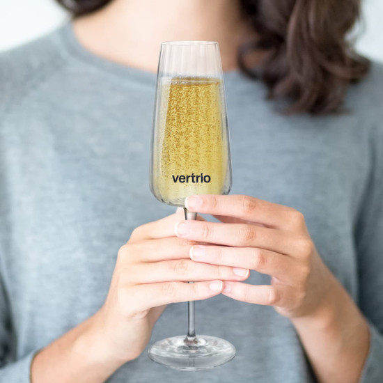 A clear champagne flute filled with bubbly, held by a person wearing a grey sweater. The glass has a logo.