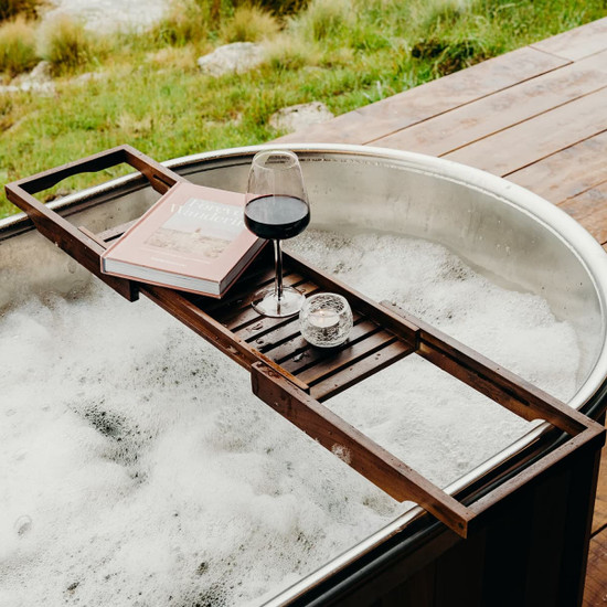 A wooden bath caddy in dark brown, holding a glass of red wine, a candle, and a book, resting over a bubbly bathtub.