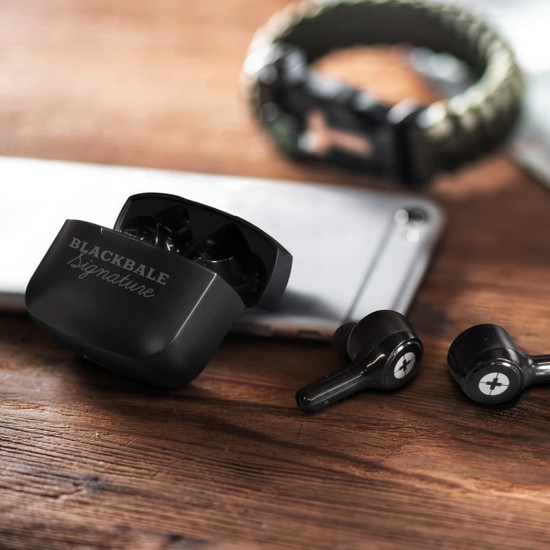 Black wireless earbuds with a charging case, placed on a wooden surface next to a smartphone and a bracelet.
