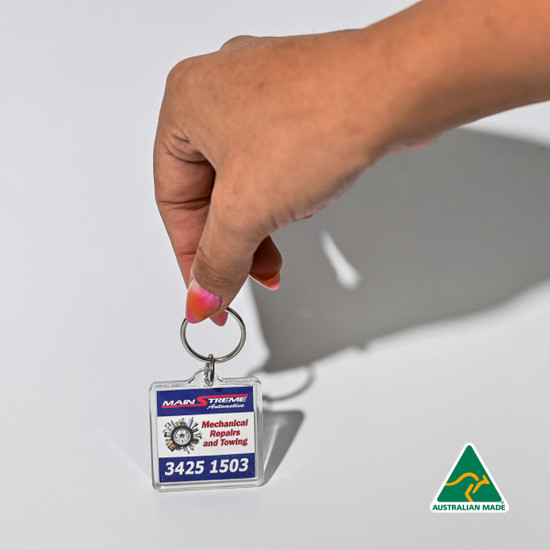 A hand holding a square acrylic keychain featuring a design in blue and white, with a logo.
