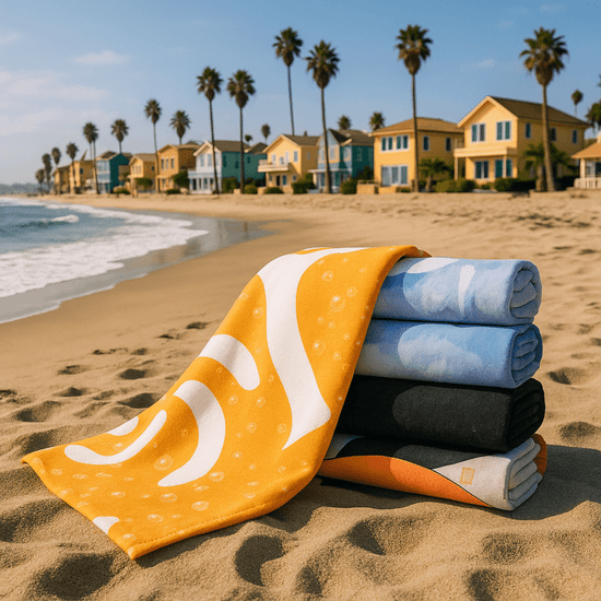 A stack of five bath towels in various colours, including yellow, blue, and black, on a sandy beach with palm trees and houses in the background.