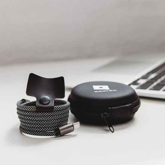 A charging cable in black and white fabric with a snap closure, packaged in a circular black case with a logo.