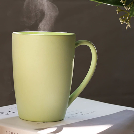 A reusable bamboo mug in a soft green colour, emitting steam, sits on a book with foliage in the background.
