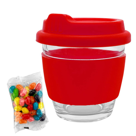 A reusable glass coffee cup with a red silicone lid and sleeve, alongside a packet of colourful jelly beans.