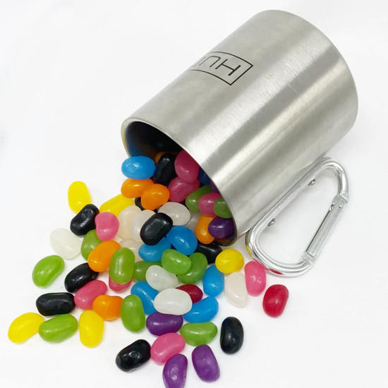 A stainless steel mug spills colourful jelly beans, including shades of yellow, pink, green, black, and white, with a carabiner.
