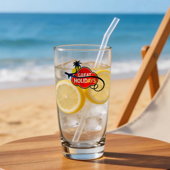 A tall glass of lemonade with lemon slices and ice, featuring a logo, sits on a wooden table by the beach.