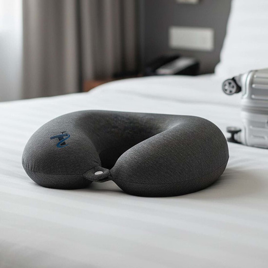 A grey neck pillow with a U-shape design rests on a neatly made bed. It has a small loop for attachment.
