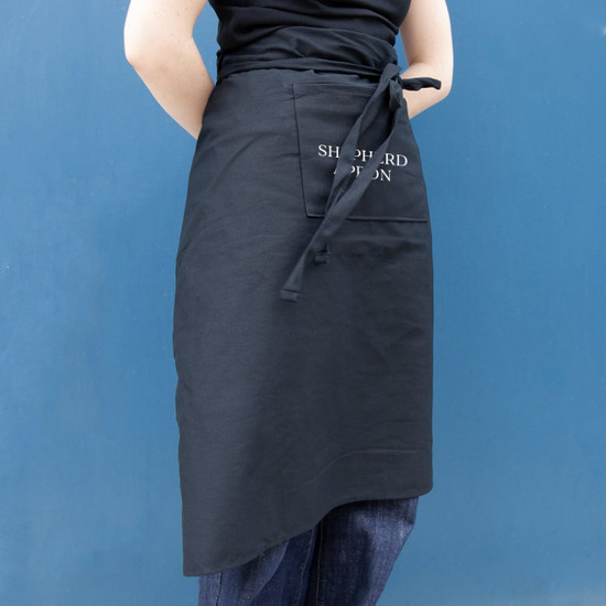 A black long waist apron with a side pocket, featuring a logo on the front.