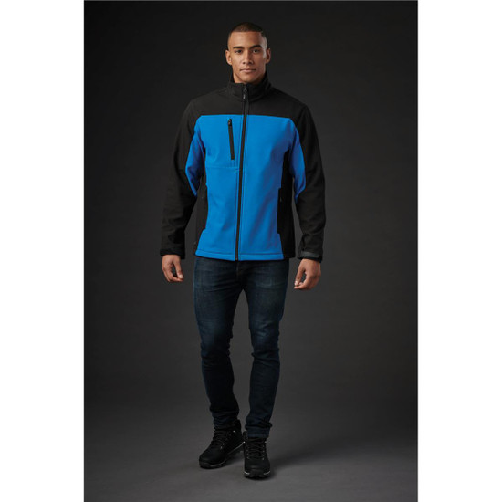 A man wearing a blue and black softshell jacket, standing against a dark background. The jacket has a zip and a logo.