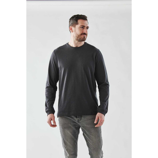 A man wearing a long sleeve tee in dark grey, paired with light grey jeans, against a plain background.
