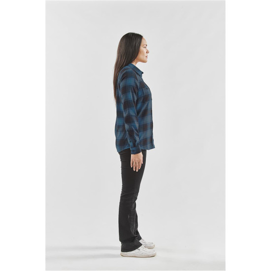 A side view of a woman wearing a blue and black checkered button-up shirt with black jeans and white shoes.