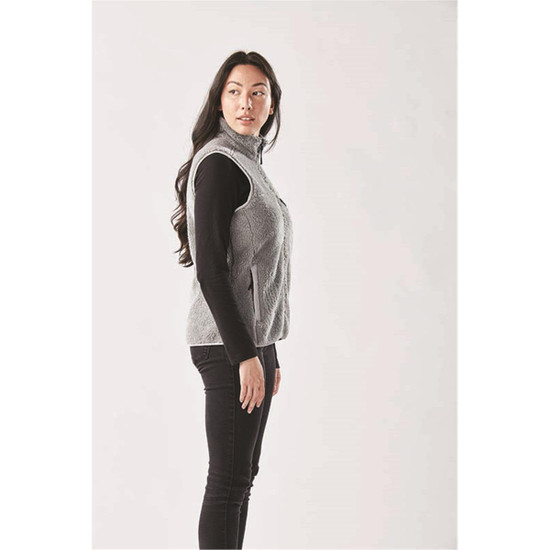 A side view of a women's grey sherpa fleece vest, worn over a black long-sleeve top, with a logo visible.