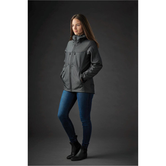 A woman stands in a dark grey jacket with pockets, paired with jeans and ankle boots, against a dark background.