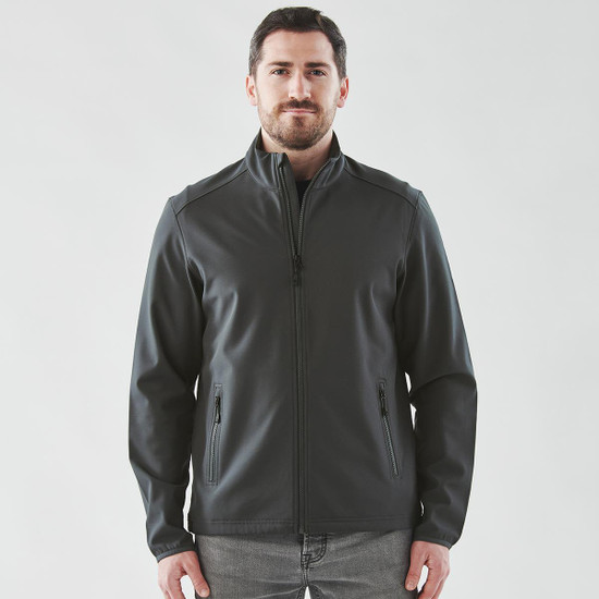 A man wearing a dark grey softshell jacket with a zip front and side pockets. The jacket has a logo.