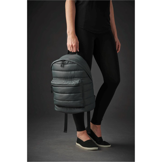 A quilted dark green backpack held by a person in black leggings and slip-on shoes, featuring a zipper and a logo.