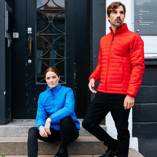 A woman in a blue quilted jacket and a man in a red quilted jacket stand on steps outside a building. Both jackets have branding.