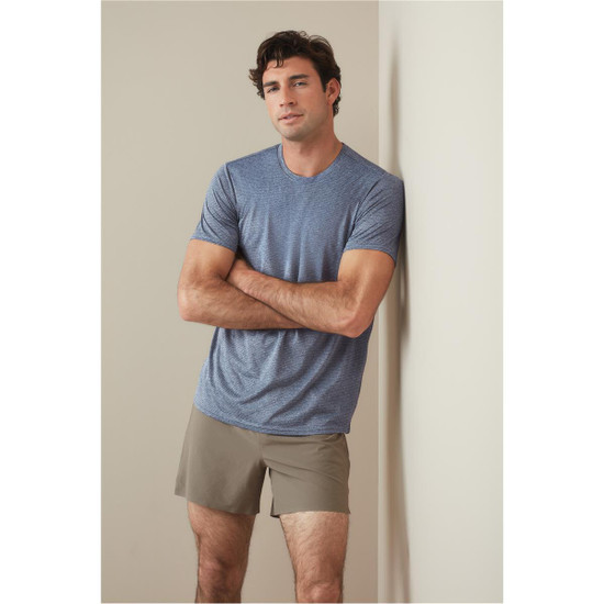 A man wearing a blue recycled sports T-shirt, leaning against a wall, with brown shorts. The shirt has a logo.