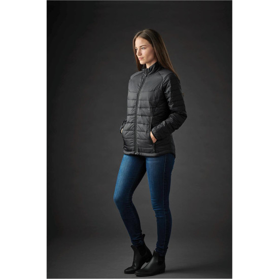 A woman wearing a black puffer jacket with a zip, paired with blue jeans and ankle boots. The jacket features a logo.