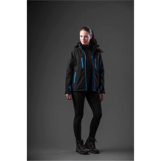 A woman in a black jacket with blue accents stands against a dark background, wearing black leggings and boots.