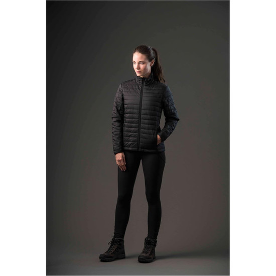 A woman models a black puffy jacket, paired with black leggings and boots, set against a dark background.